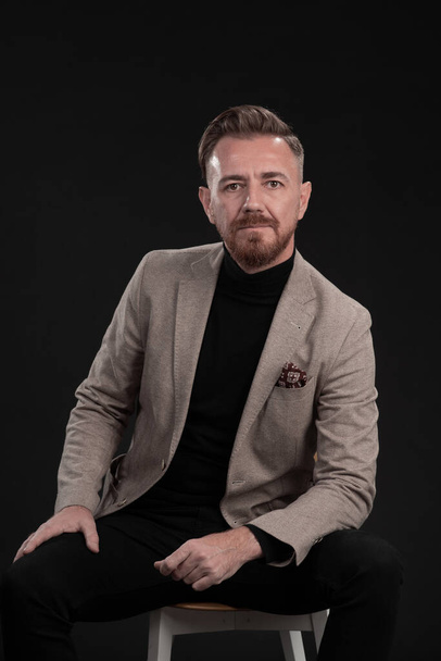 Portrait of adult businessman wearing trendy suit and sitting in modern studio on stylish chair against the black background - Фото, зображення