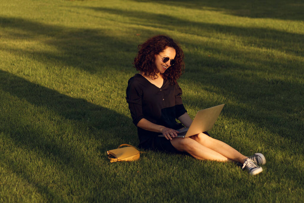 Mujer bastante joven en gafas de sol sentado en el césped con piernas durante el día de verano, mientras que el uso de la computadora portátil. - Foto, Imagen