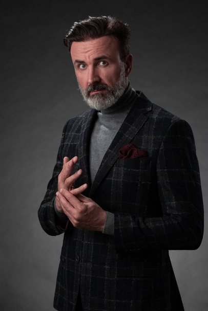 Portrait of a stylish elegant senior businessman with a beard and casual business clothes in photo studio isolated on dark background gesturing with hands - Фото, зображення