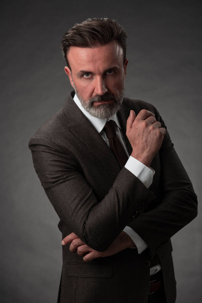 Portrait of a stylish elegant senior businessman with a beard and casual business clothes in photo studio isolated on dark background gesturing with hands - Fotó, kép