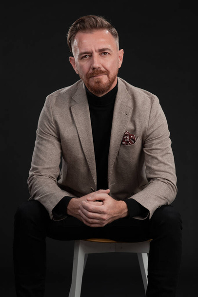 Portrait of adult businessman wearing trendy suit and sitting in modern studio on stylish chair against the black background - Foto, afbeelding