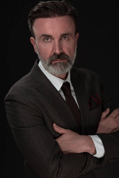 Portrait of a stylish elegant senior businessman with a beard and casual business clothes in photo studio isolated on dark background gesturing with hands - Foto, imagen