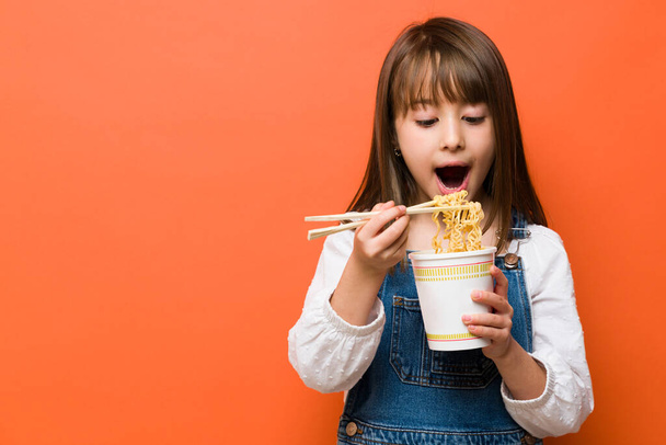 Pretty brunette little girl opening her mouth and eating some ramen noodles from a cup using chopsticks - Valokuva, kuva