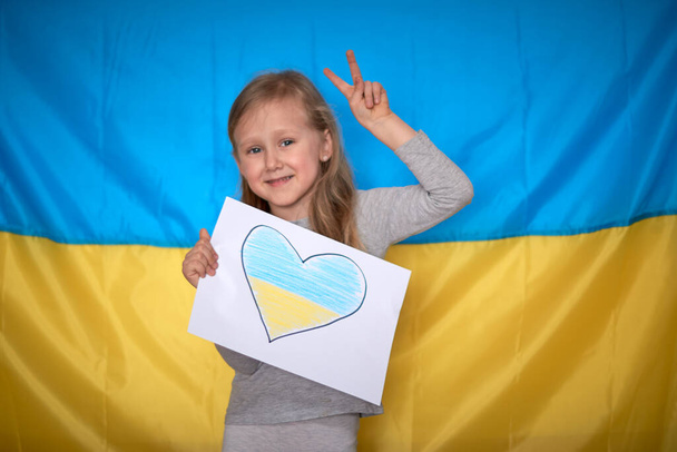 Girl hands holding paper banner with drawn yellow-blue heart on Ukrainian flag background. Kids support Ukraine, stop war, stay with Ukraine hashtag - Photo, Image