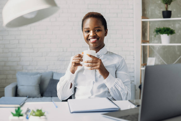 Retrato interior de la mujer afroamericana complacida mirando a la cámara mientras posaba en la oficina durante el descanso del café. Retrato de mujer de negocios. Elegante negro americano - Foto, imagen