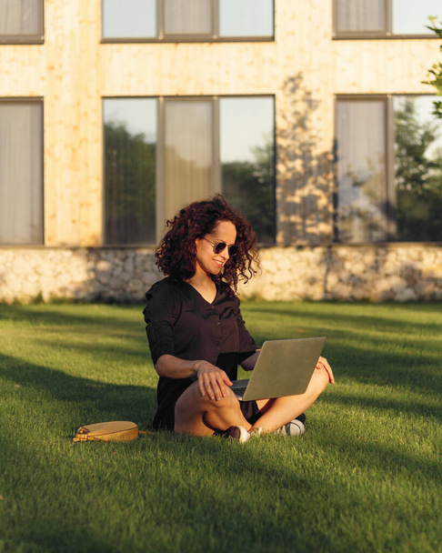 Allegro riccio giovane donna in occhiali da sole seduto su erba verde con le gambe incrociate durante la giornata estiva durante l'utilizzo di laptop e parlare in chat. - Foto, immagini