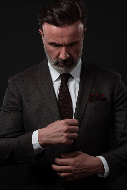 Portrait of a stylish elegant senior businessman with a beard and casual business clothes in photo studio adjusting suit - Fotó, kép
