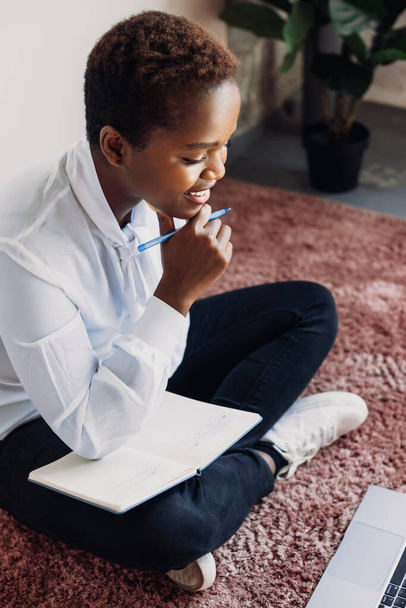 Étudiant afro-américain tenant une classe d'apprentissage de cahier en utilisant regarder des leçons de tutorat en ligne tout en étant assis sur le sol à la maison. Devoir. Moderne - Photo, image