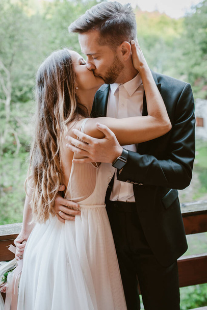 Νύφη και γαμπρός φιλιούνται ενώ στέκονται στο μπαλκόνι ενός ξύλινου σπιτιού στο δάσος. Κοντινό πορτραίτο. Εκπληκτική καλοκαιρινή φύση. - Φωτογραφία, εικόνα