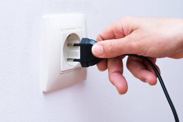 Plugging a black electrical plug with a womans hand into a white plastic socket on a wall. - Foto, Imagem