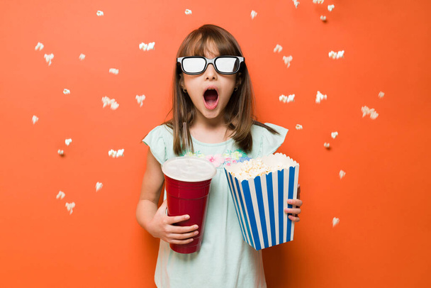 Cute girl wearing 3d glasses and looking shocked while watching a movie in the cinema theater with popcorn flying in the air - Fotoğraf, Görsel