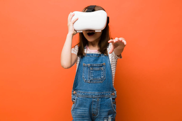 Portret van een mooi klein meisje met VR-bril en verbaasd kijkend naar haar virtuele ervaring - Foto, afbeelding