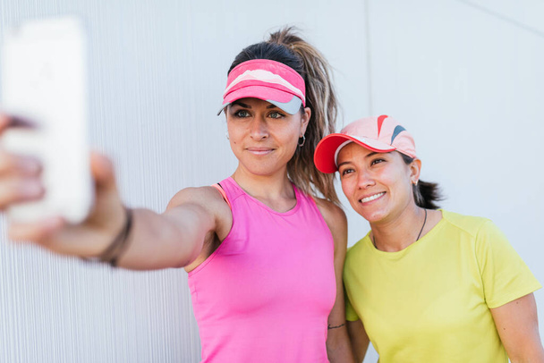 Dos mujeres en forma usando ropa deportiva tomando fotos de selfie mientras hacen ejercicio en la ciudad. Dos mujeres deportivas sonriendo y haciendo selfie después de correr. - Foto, Imagen