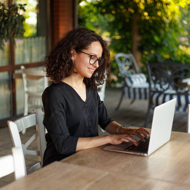 Terasta otururken dizüstü bilgisayarda çalışan neşeli genç bir kadının yan görüntüsü. - Fotoğraf, Görsel