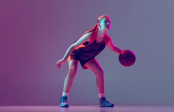 Portrait of young girl, teen, basketball player in motion, training isolated over gradient pink purple background in neon. Concentration - 写真・画像