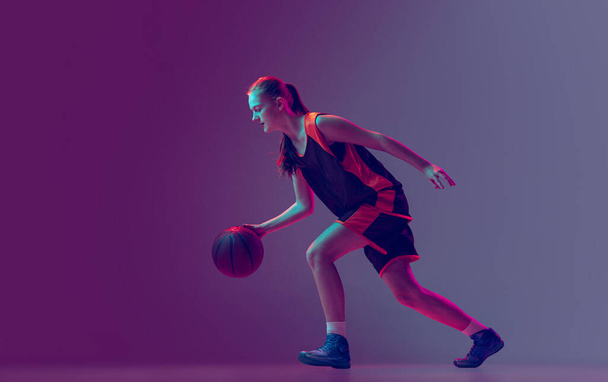 Portrait of young girl, teen, basketball player in motion, training isolated over gradient pink purple background in neon. Side view - Foto, Bild