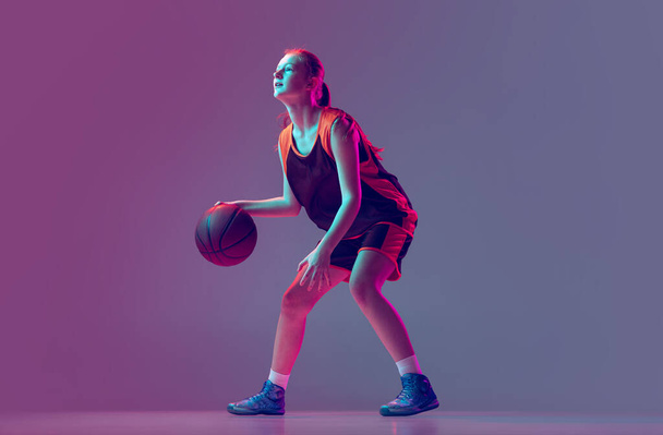Portrét mladé dívky, teen, basketbalista v pohybu, trénink izolovaný přes gradient růžové fialové pozadí v neonu - Fotografie, Obrázek