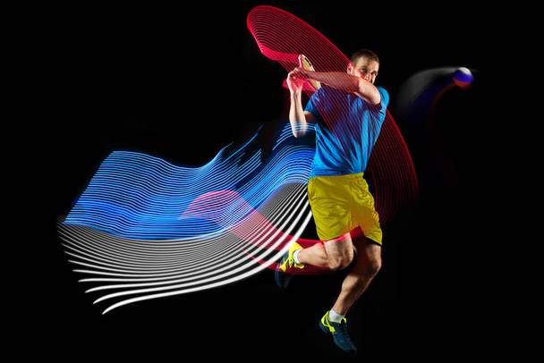 Portrait of young man playing tennis isolated on dark background with mixed colored stipes. Concept of sport, hobby, challenges, peace, politics - Photo, Image