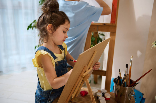 Taideluokka Suloinen valkoihoinen lapsimaalaus kankaalle. Kaunis tyttö keltaisessa t-paidassa nauttimassa luovasta taiteesta kotona. Harrastus, taide, luovuus ja lasten viihde käsite - Valokuva, kuva