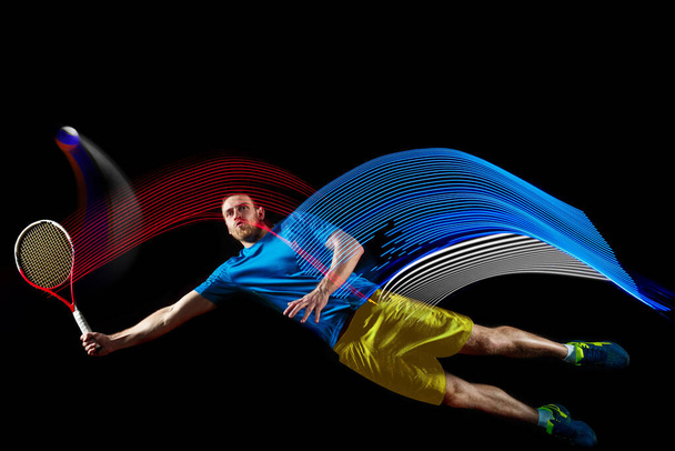 Portrait of young man playing tennis isolated on dark background with mixed colored stipes. Concept of sport, hobby, challenges, peace, politics - Zdjęcie, obraz