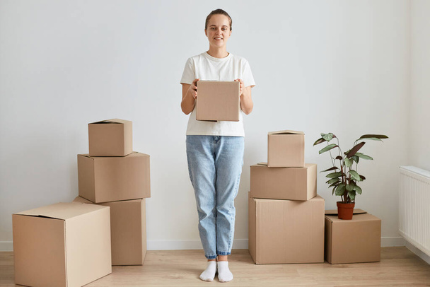 Full length portrait of woman wearing white t shirt standing near cardboard boxes with belongings, holding parcel with personal pile, looking at camera with cute smile. - Photo, Image