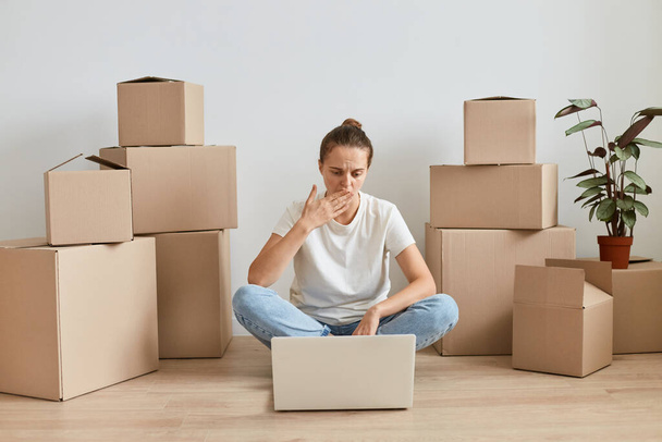 Indoor shot of woman wearing white t shirt sitting on floor surrounded with cardboard boxes with belongings and working on laptop, looking at screen, covering mouth, having mistake. - Фото, изображение