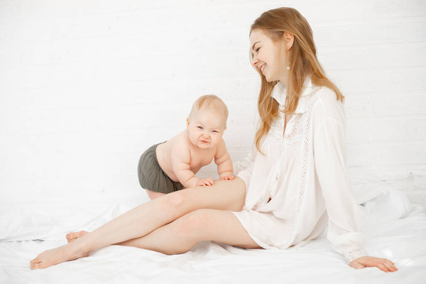 Młody boso uśmiechnięta kobieta w koszuli z nagim dzieckiem w pieluszce siedzi w białej sypialni. Rozwój dziecka, relacje z mamą - Zdjęcie, obraz