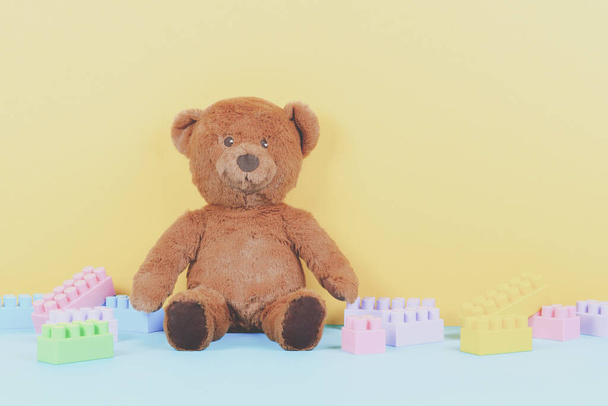 Baby kids toys background with brown teddy bear and colorful building blocks bricks on pastel yellow and blue background. Front view - Photo, Image