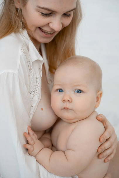 Vertical photo of smiling blond gentle woman with bare chest, holding little baby eating, breastfeeding before sleep - Foto, afbeelding