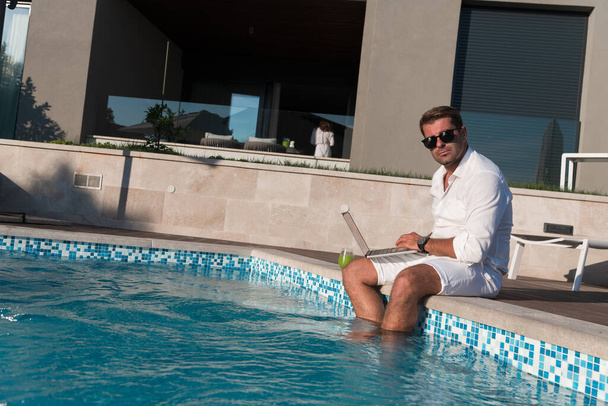 Een moderne oudere man geniet van het zwembad tijdens het werken op zijn laptop naast een modern luxe huis. Selectieve focus  - Foto, afbeelding
