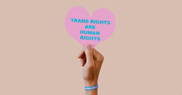 um jovem caucasiano, usando uma pulseira padronizada com a bandeira do orgulho transgênero, tem um coração rosa com o texto direitos transgêneros são direitos humanos recortados nele, em um fundo rosa pálido. - Foto, Imagem