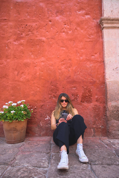 Orta çağ caddesinde kırmızı bir duvarda otururken gülümseyen ve cep telefonunu kontrol eden beyaz bir kadın. Dikey. Kopyalama alanı ile. - Fotoğraf, Görsel