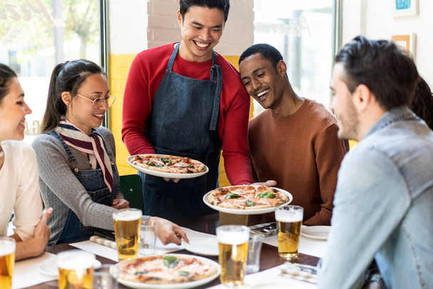 Zaufany kelner serwujący pyszne pizze margherita wielokulturowym przyjaciołom w przytulnej pizzerii - wieloetniczni przyjaciele bawiący się razem w pizzerii jedząc pizzę i pijąc blond piwo - Zdjęcie, obraz