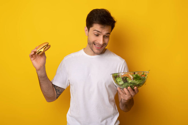 Retrato de un joven divertido sosteniendo hamburguesa y ensalada - Foto, imagen