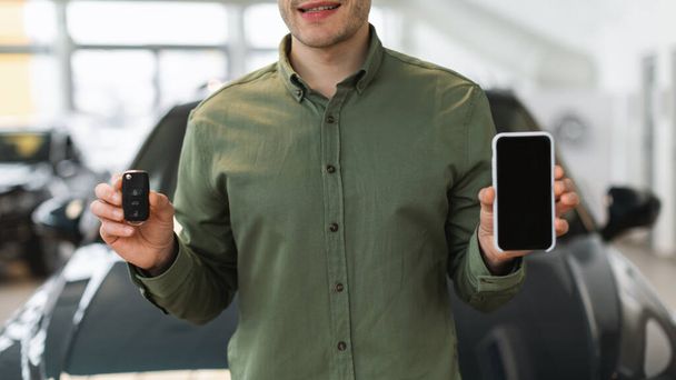 Onherkenbare jongeman toont nieuwe autosleutel en smartphone met blanco scherm bij auto-dealer, mockup - Foto, afbeelding