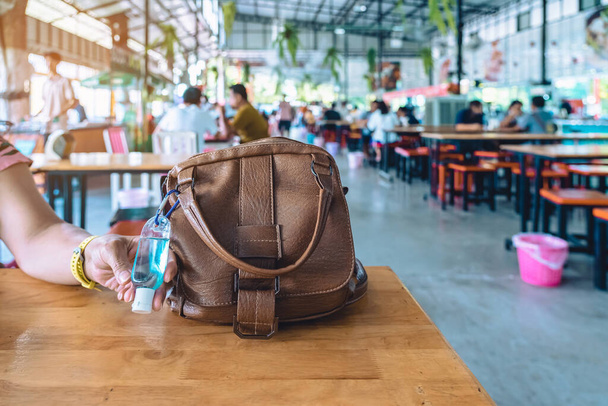 Mini portable blue alcohol gel bottle to kill Corona Virus(Covid-19) hang on brown leather shoulder bag of a woman on table at cafeteria. New normal lifestyle. Selective focus on alcohol gel. - Foto, Imagen