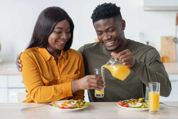 Giovani sposi neri felici che fanno colazione insieme, marito che versa succo di frutta dalla brocca - Foto, immagini