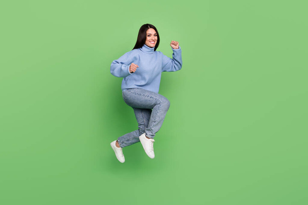 Photo of lucky pretty lady dressed blue sweater rising fists jumping high running fast isolated green color background - Φωτογραφία, εικόνα