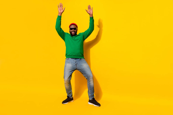 Photo of positive man dance raise hands wear sunglass red beanie green shirt jeans isolated yellow color background - Valokuva, kuva