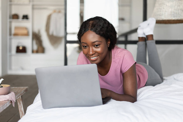 Feliz Milenial Negro Femenino Usando el ordenador portátil acostado en la cama interior - Foto, Imagen