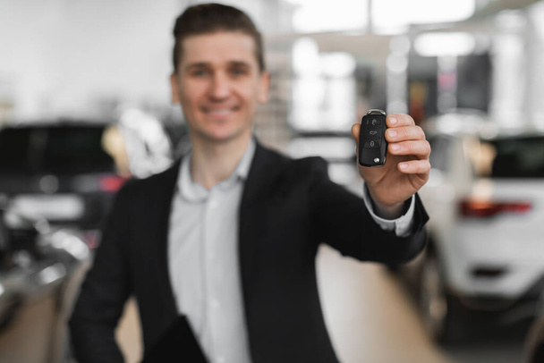 Selbstbewusster junger Verkäufer zeigt Autoschlüssel vor Kamera in modernem Autohaus, selektiver Fokus - Foto, Bild