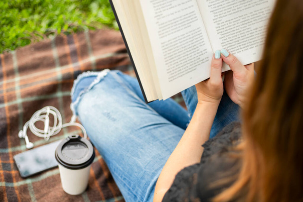 公園の毛布の上に座っている間、コーヒー読書本のカップを持つジーンズの女性. - 写真・画像