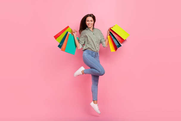 Photo of excited lucky woman wear striped shirt rising shopping bags jumping high isolated pink color background - Photo, Image