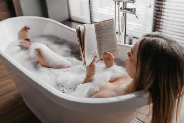 Journée détente et spa. Femme insouciante couchée dans un bain mousseux, lisant un livre et appréciant une histoire intéressante - Photo, image
