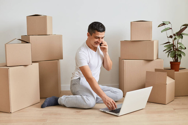 Indoor shot of man wearing white t shirt sitting on floor surrounded with cardboard boxes with belongings and working on laptop and talking phone, works in delivery service online. - Foto, Imagem