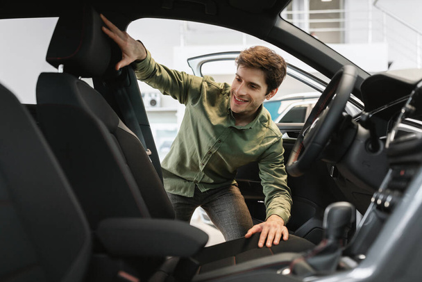 Feliz joven mirando dentro del salón de coches, la selección de automóviles nuevos en la concesionaria moderna - Foto, Imagen