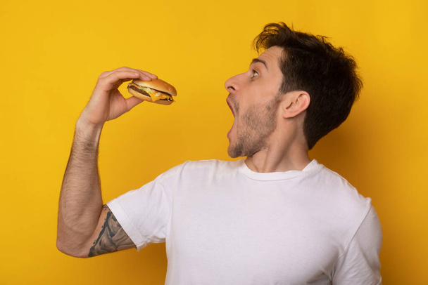 Divertido chico sosteniendo hamburguesa mordiendo sándwich en el estudio - Foto, imagen
