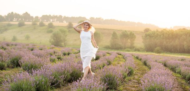 Beautiful young woman wearing a white dress walking in the middle of a lavender field in bloom - Φωτογραφία, εικόνα