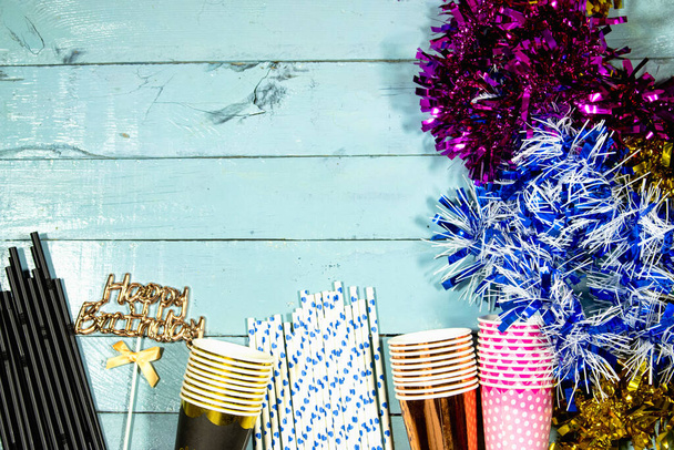 誕生日パーティーのコンセプト使い捨てカップとストローと青の木製テーブルの上にカラフルなティンセル  - 写真・画像