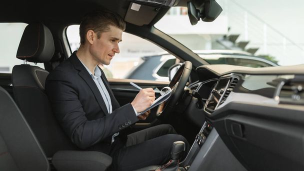 Joven vendedor de automóviles sentado dentro de un coche nuevo, haciendo chequeo, anotando datos en un concesionario moderno, panorama - Foto, imagen